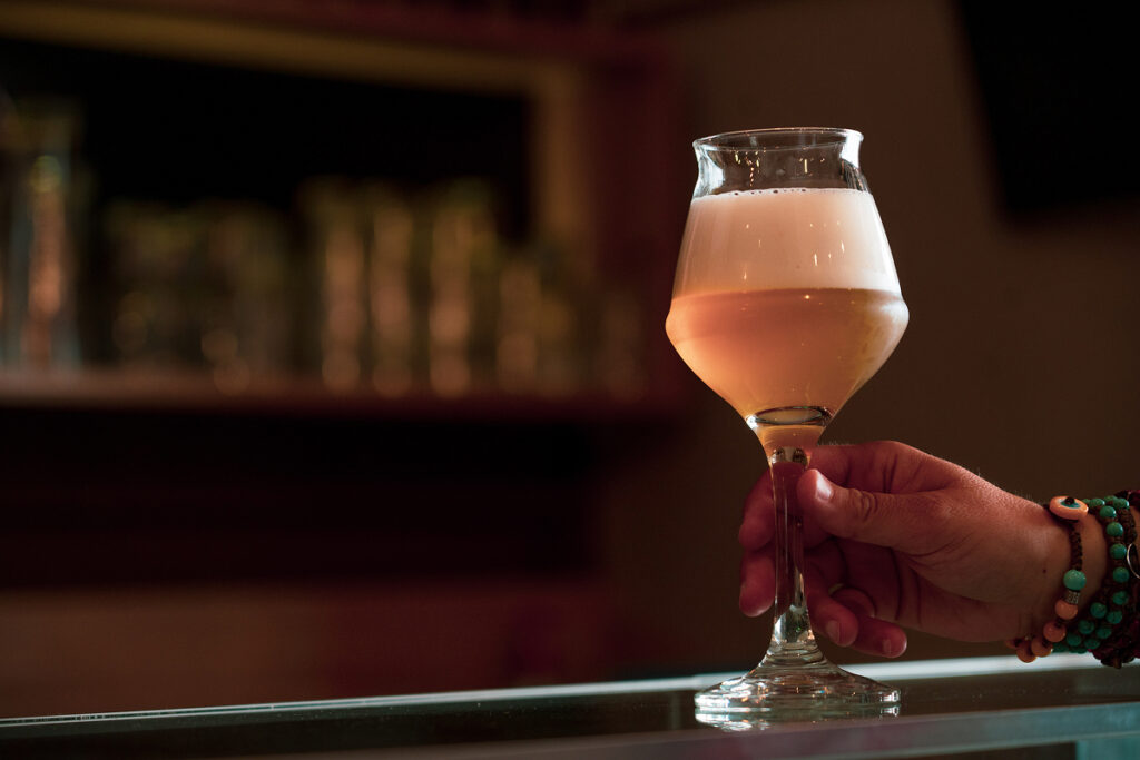 Calice di birra chiara senza glutine tenuto da una mano su un bancone.